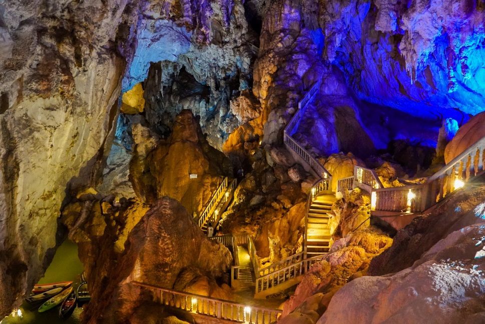 Un escalier en bois serpente à travers une grande grotte avec des formations rocheuses colorées illuminées par des lumières bleues et jaunes, avec des bateaux sur l'eau en contrebas.