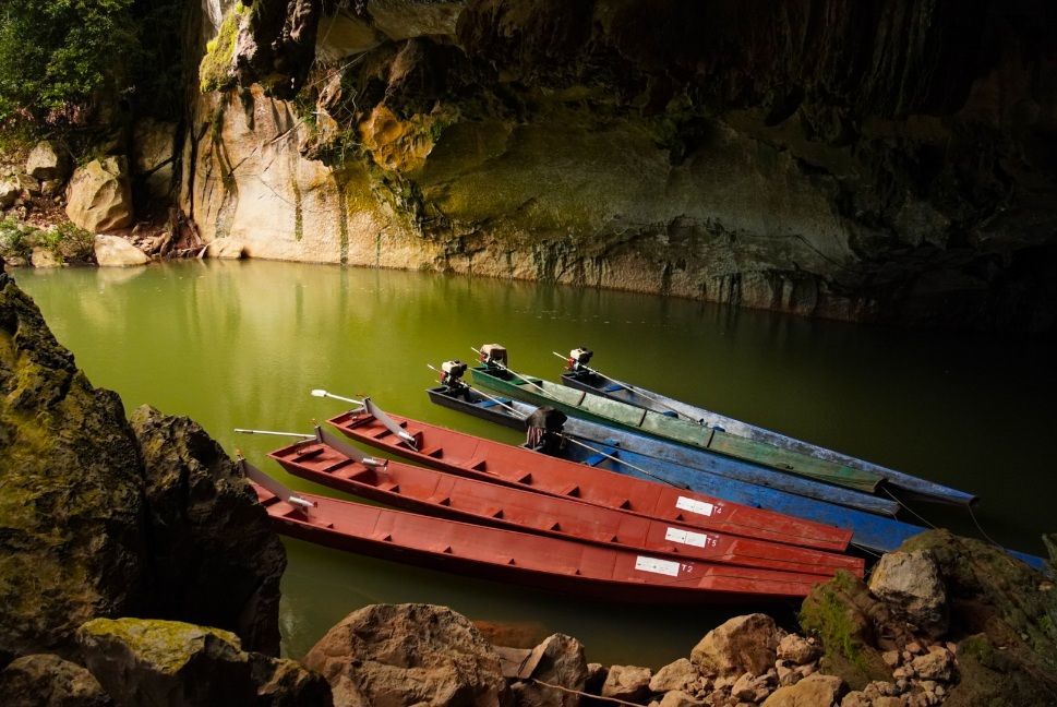 Plusieurs bateaux à longue queue rouges, bleus et verts sont amarrés sur une eau vert trouble, à l'intérieur d'une grotte rocheuse.