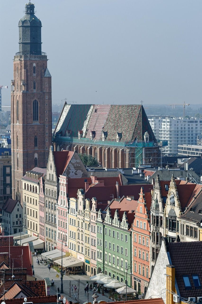 Una vista aérea de una plaza de ciudad europea con coloridos edificios históricos y una gran torre de iglesia de ladrillo al fondo.