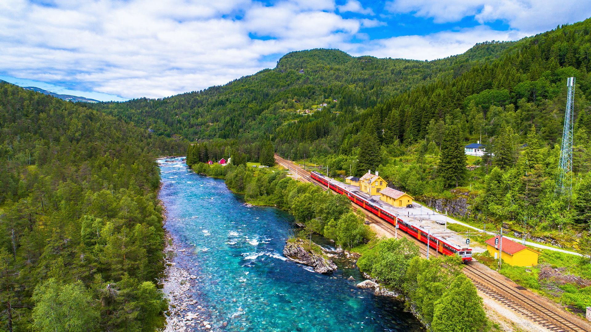 Una veduta aerea di un treno rosso in stazione, costeggiando un fiume turchese che attraversa una valle montana lussureggiante e boscosa.