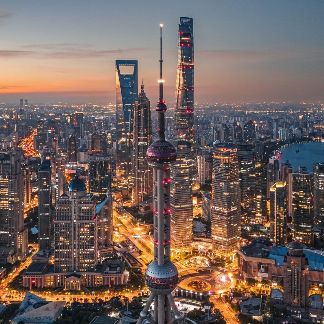 Lo skyline illuminato di una città moderna con numerosi grattacieli e un'imponente torre, vista dall'alto al crepuscolo.