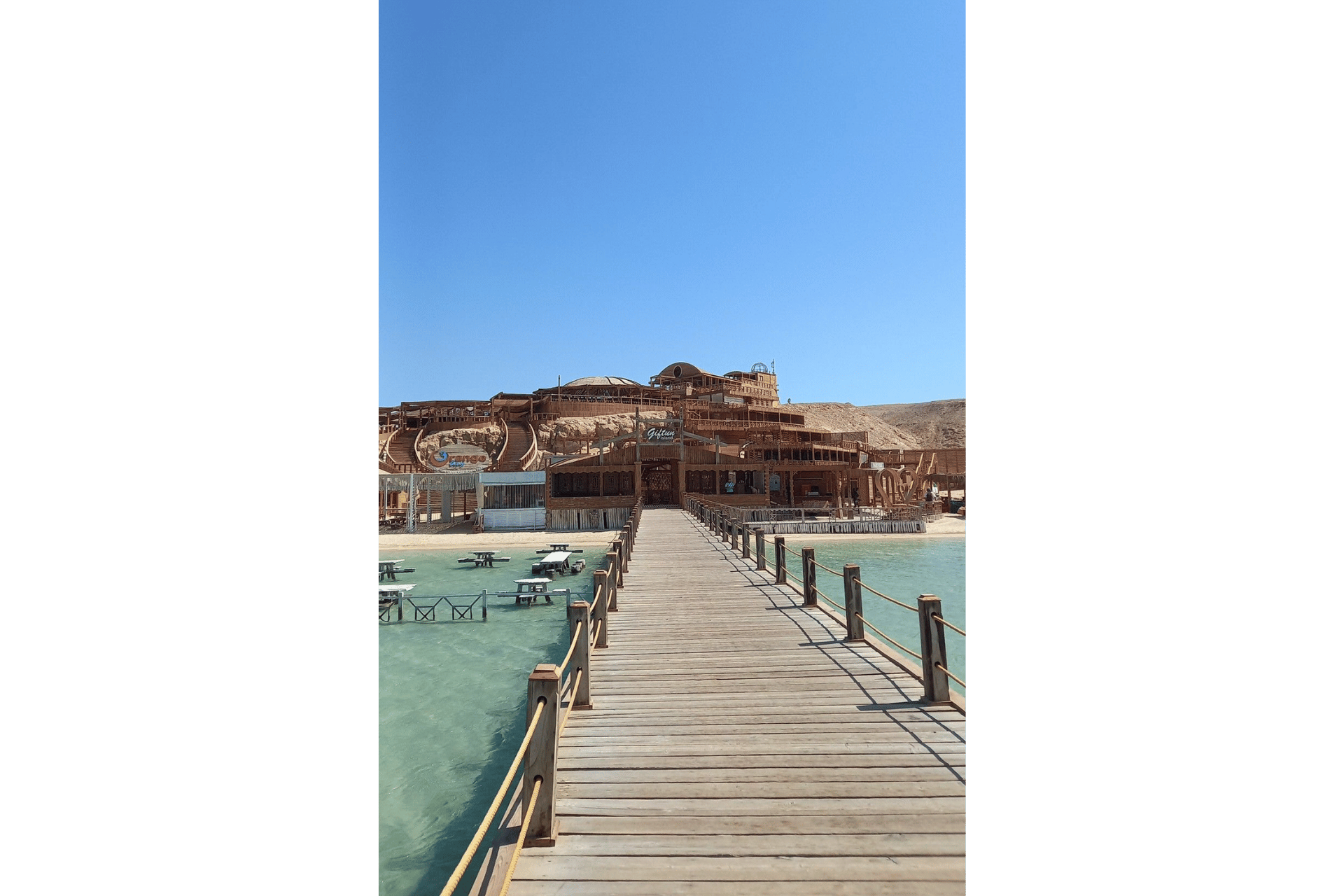 Un lungo molo di legno si estende su acque turchesi e limpide verso un grande resort balneare in legno costruito su un versante sabbioso sotto un cielo azzurro e sereno.