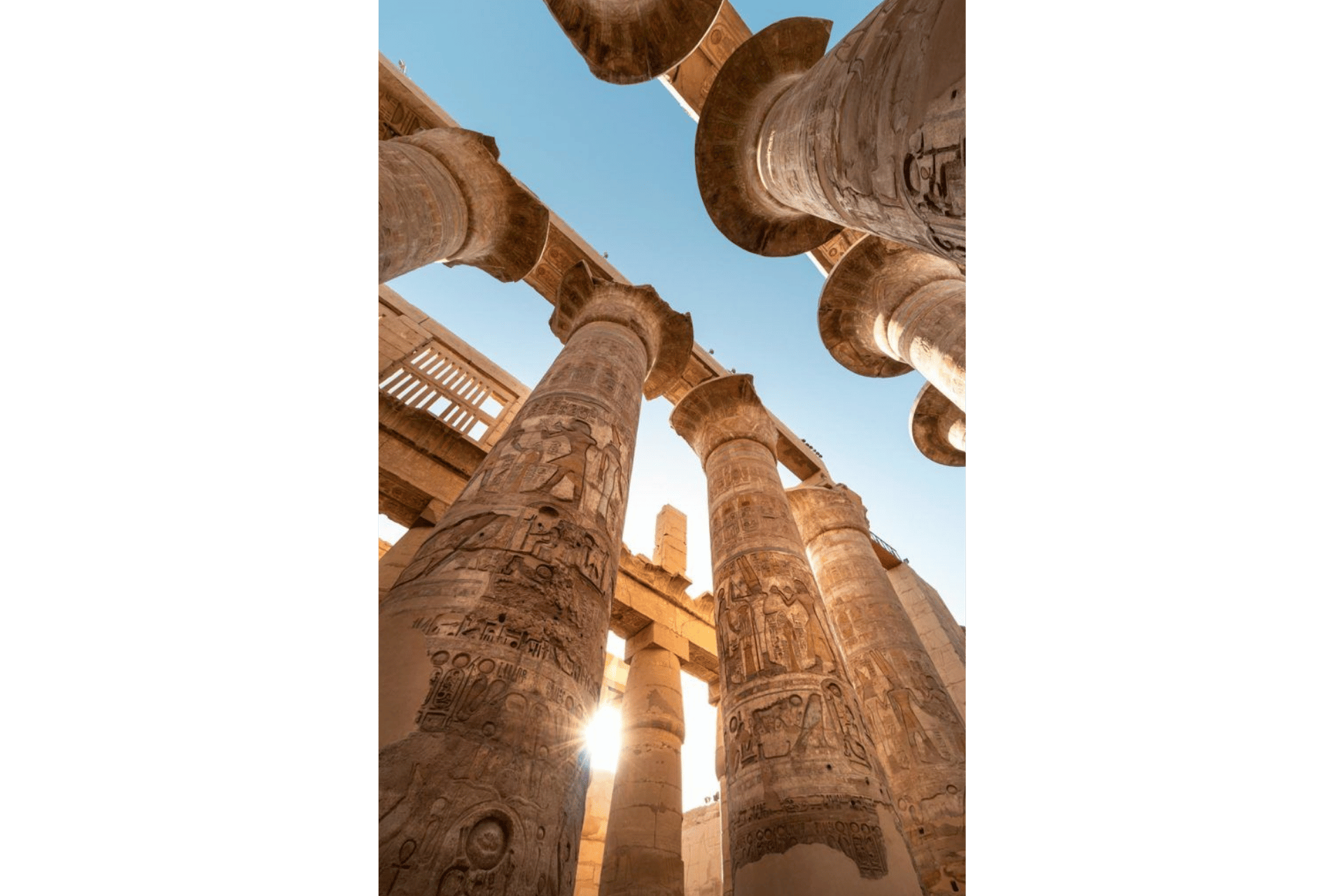 Vista dal basso di imponenti colonne di pietra con geroglifici egizi, con il sole che splende in un cielo azzurro e limpido.