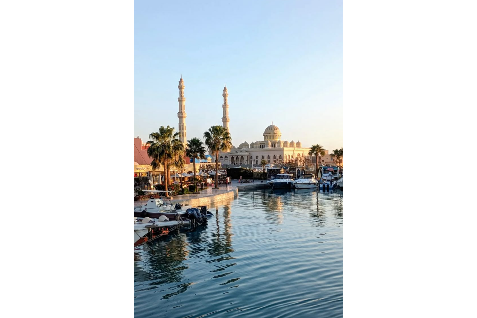Una grande moschea con due minareti si affaccia su un porto turistico, pieno di barche ormeggiate e palme, sotto un cielo sereno.