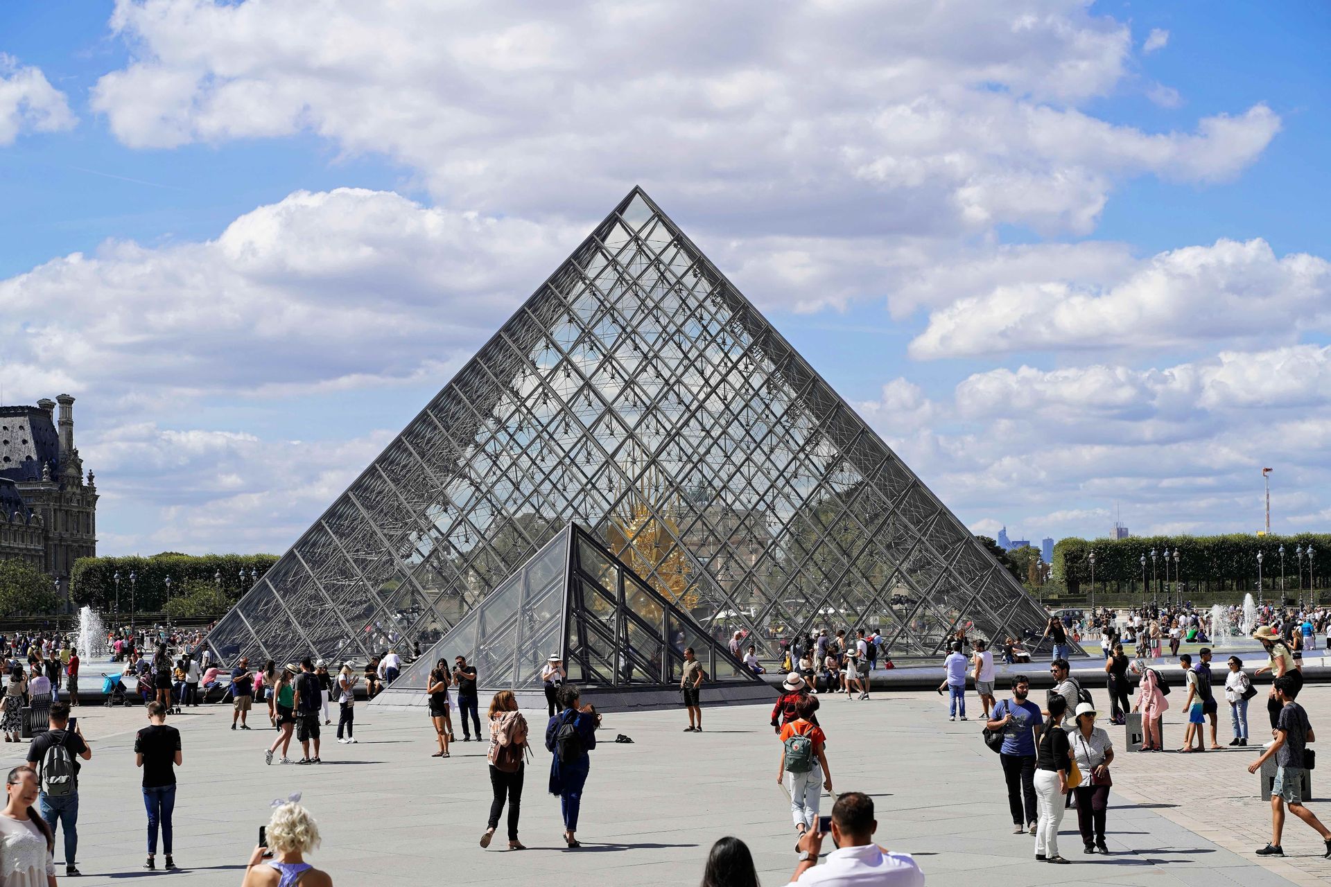 Una grande folla di persone passeggia attorno a una piramide di vetro in un ampio cortile pavimentato sotto un cielo parzialmente nuvoloso.