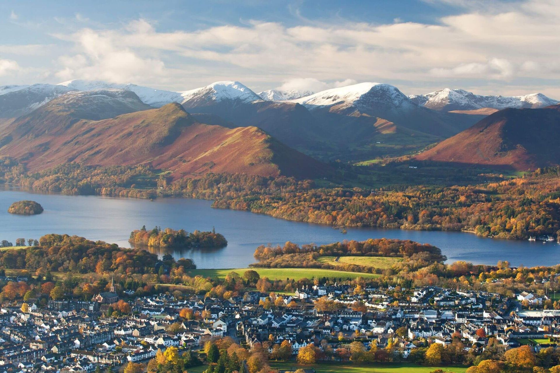 A high-angle view of a town on the shore of a large lake, with autumn-colored forests and snow-capped mountains behind it.
