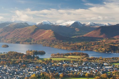 An aerial view of a lakeside town in autumn, with a backdrop of large, snow-capped mountains under a partly cloudy sky.