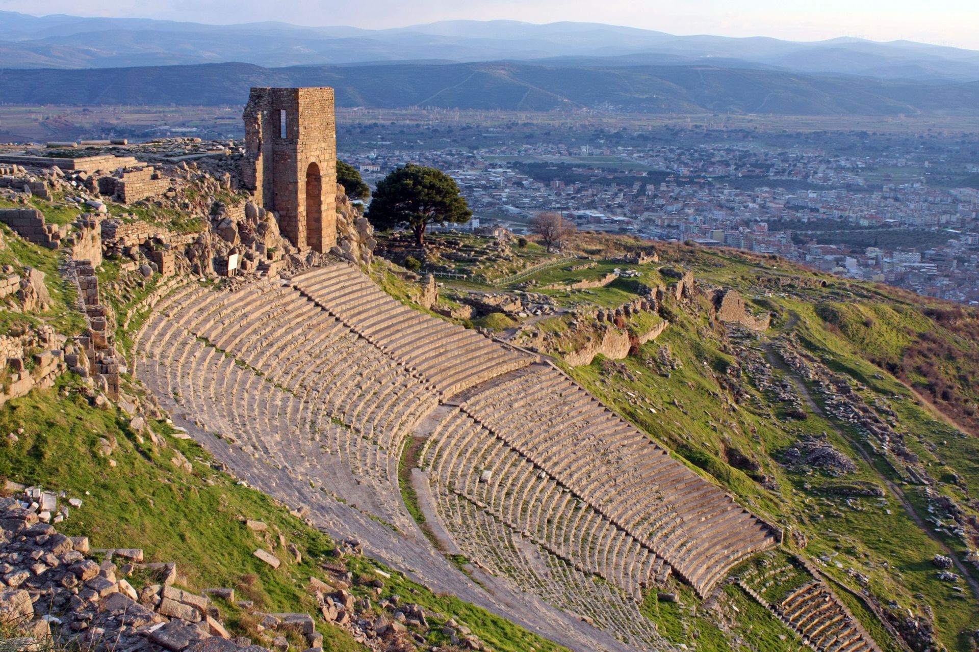 An ancient stone amphitheater built into a grassy hillside, with ruins and a modern city in the background.