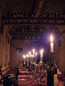 Una larga mesa de comedor en una bodega de vinos rústica, iluminada por numerosas velas en botellas de vino y con estantes llenos de botellas al fondo.