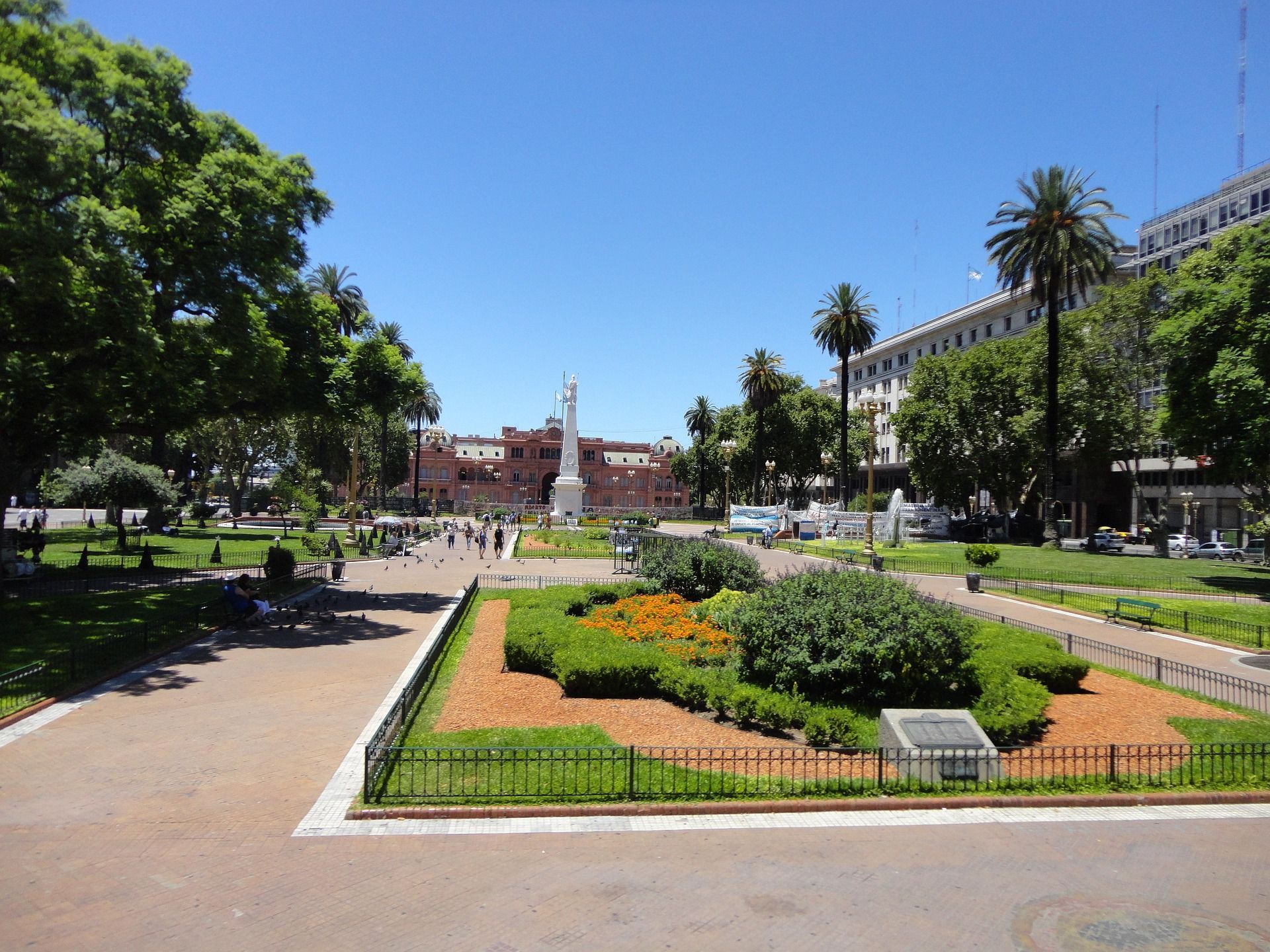 Un grande parco cittadino in una giornata di sole, con viali pavimentati, un monumento bianco e un lontano palazzo rosa circondato da alberi.