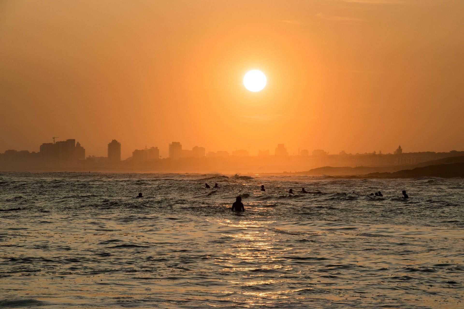 Sagome di surfisti nell'oceano durante un tramonto dorato, con lo skyline di una città visibile all'orizzonte.