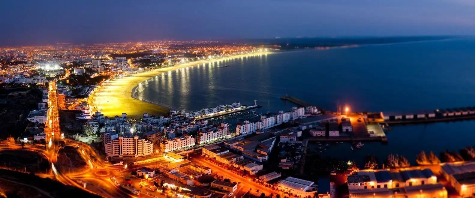 Vue aérienne nocturne d'une ville côtière lumineuse, avec des lumières éclairant une longue plage incurvée et un port.