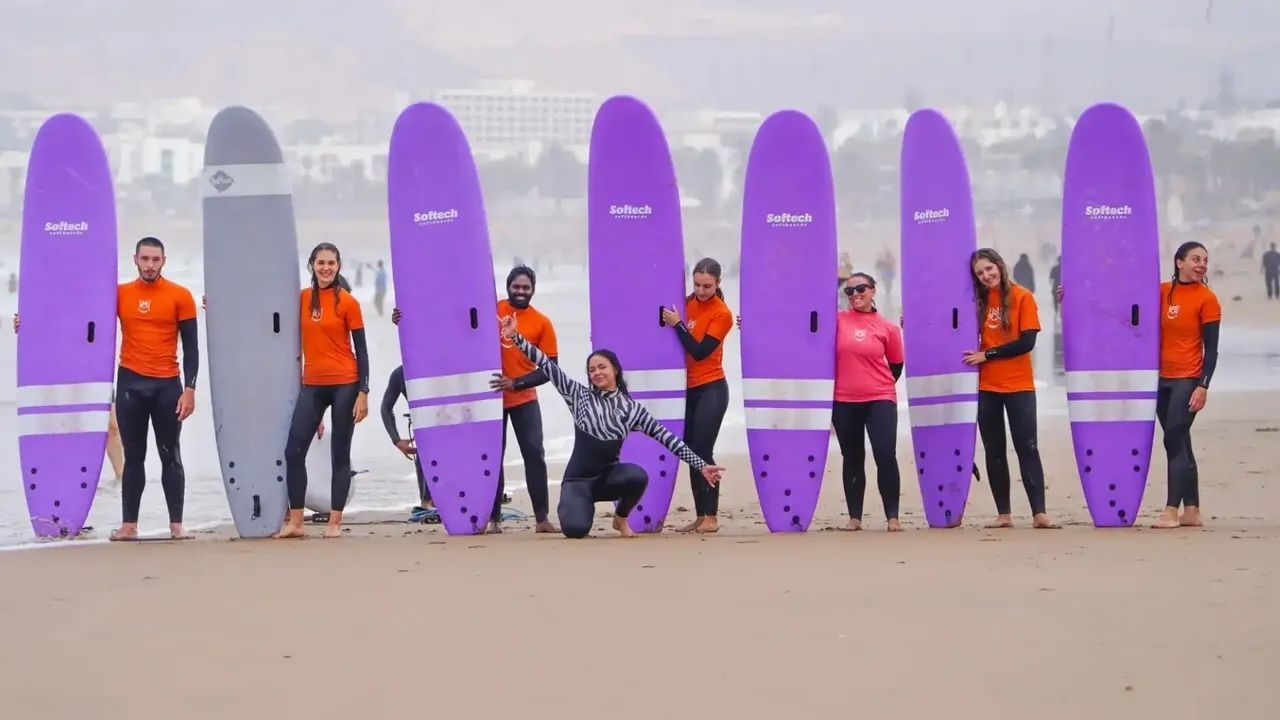Un groupe WeRoad en voyage posant avec leurs planches de surf violettes et grises sur une plage de sable avant une leçon de surf.