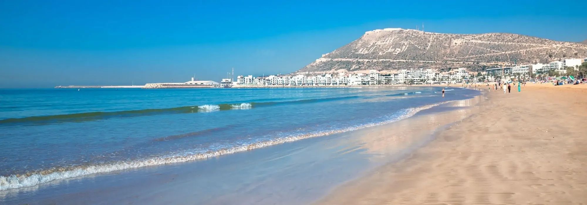 Vue panoramique d'une vaste plage de sable fin où de douces vagues caressent le rivage, avec une ville côtière et une grande colline en arrière-plan sous un ciel bleu dégagé.