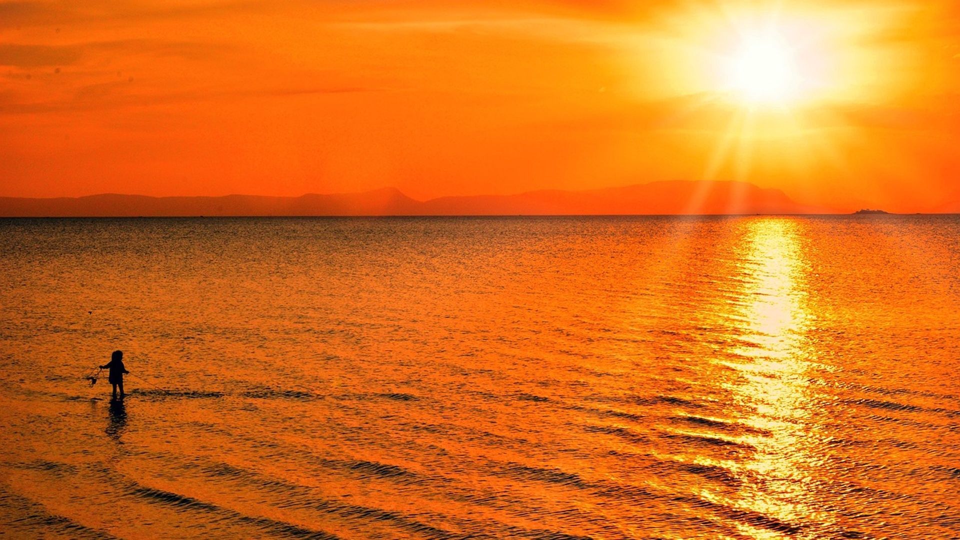 Une silhouette d'une personne debout dans la mer peu profonde au coucher de soleil orange éclatant, le soleil se reflétant sur l'eau.