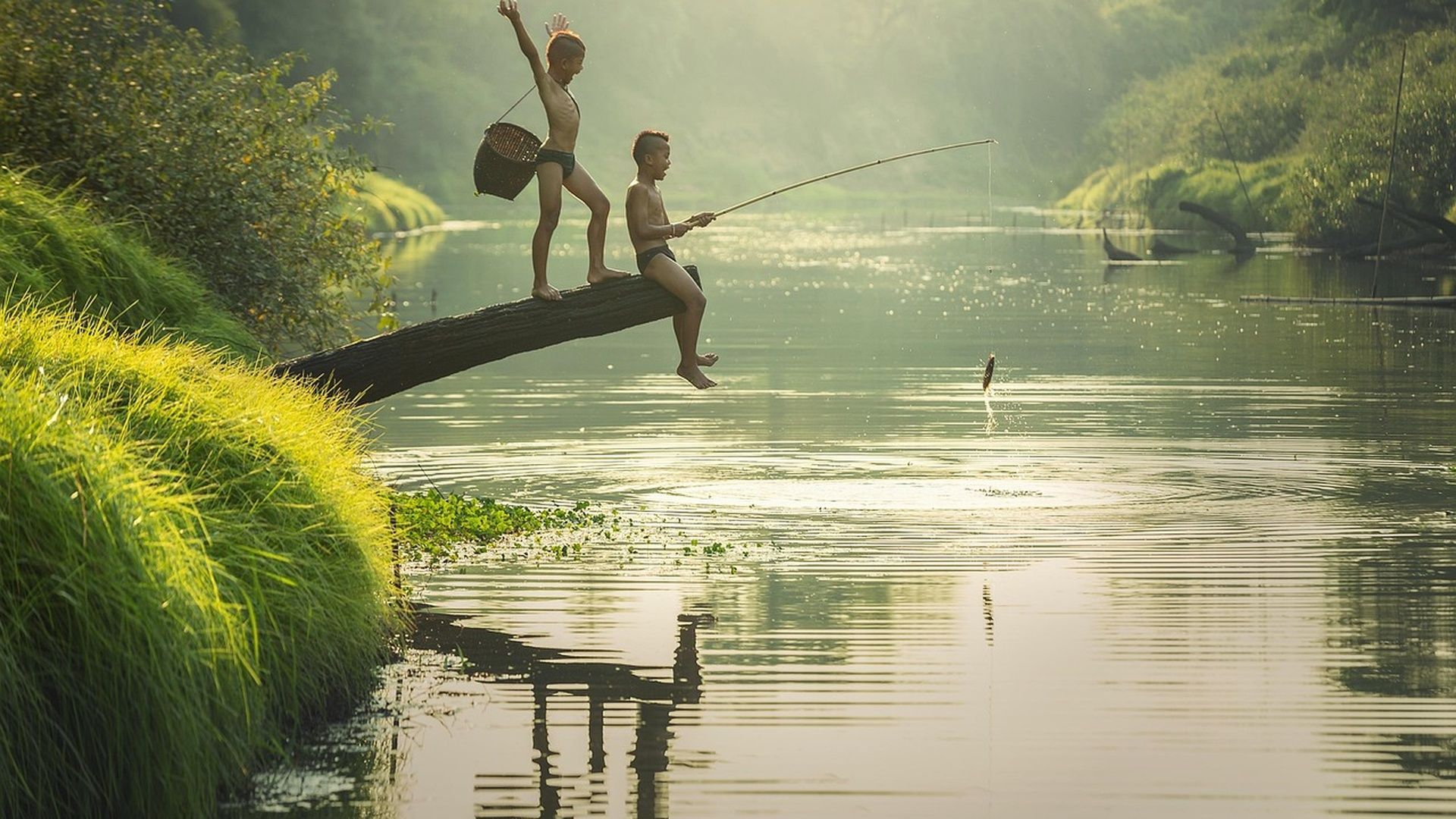 Deux garçons sur une bûche enjambant une rivière : l'un pêche à la canne et attrape un poisson bondissant, tandis que l'autre célèbre derrière lui avec un panier.