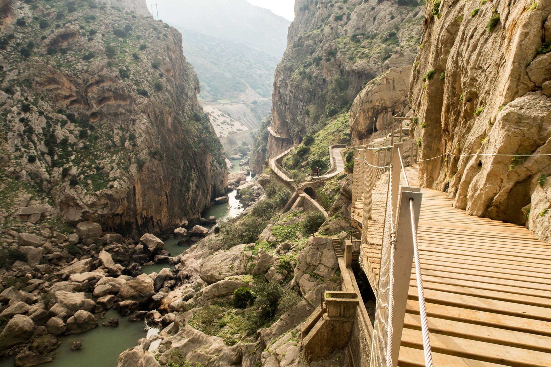 A narrow wooden boardwalk snakes along a sheer rock cliff face high above a river in a deep canyon.