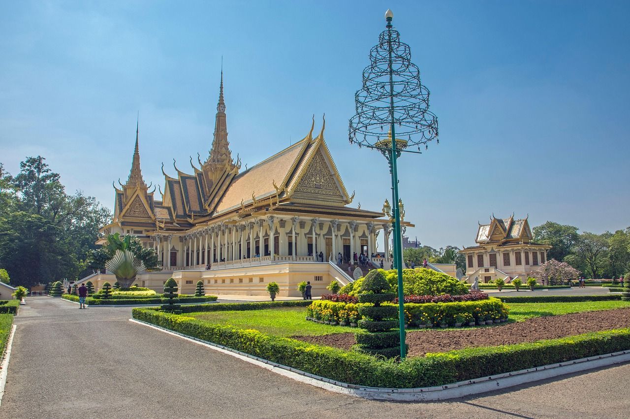 Un palais orné avec un toit doré et des flèches se dresse derrière un jardin bien entretenu doté d'un chemin pavé, sous un ciel bleu clair.