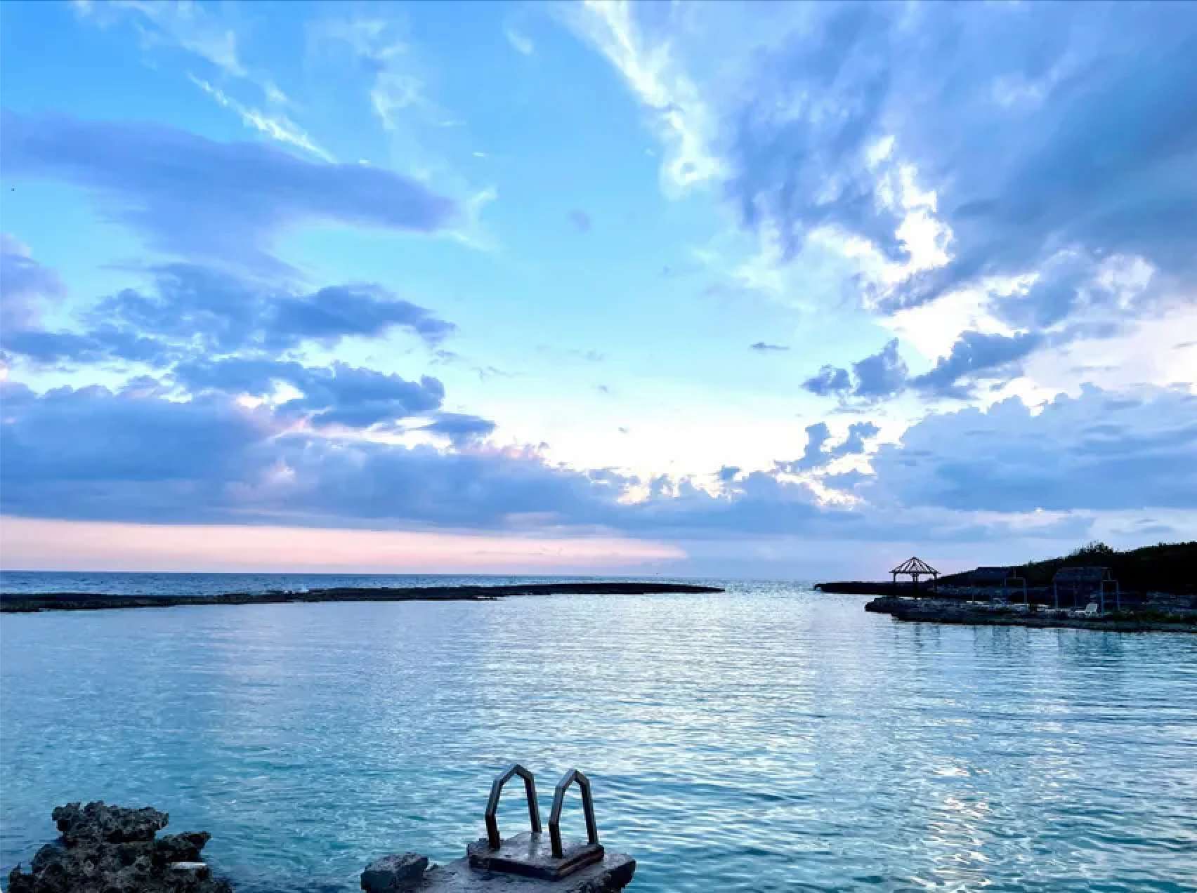 Eine Metallleiter führt in eine ruhige, klare blaue Bucht, umgeben von Felsen unter einem bewölkten Himmel in der Dämmerung.