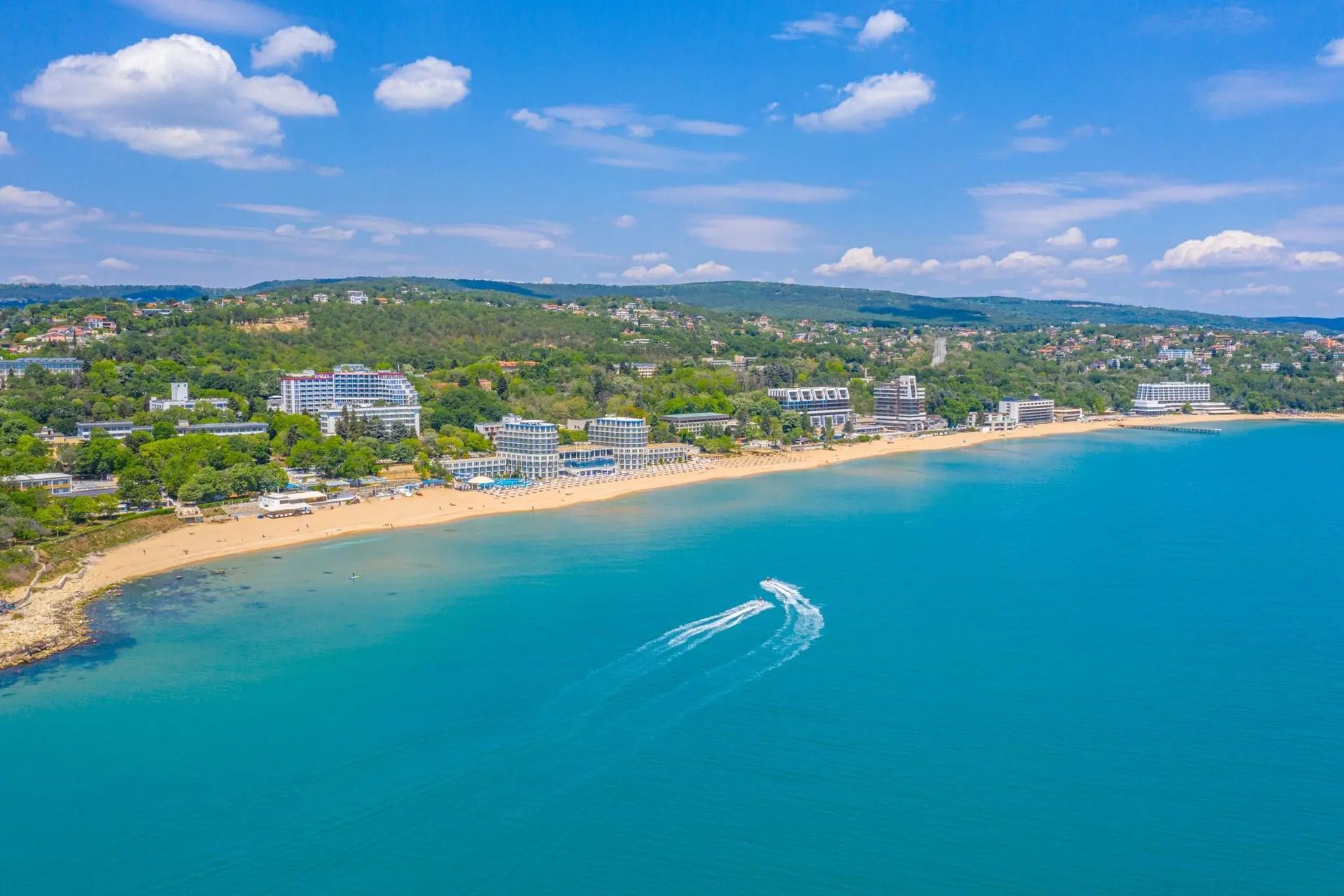 Vista aerea di due moto d'acqua su acque turchesi, vicino a una spiaggia sabbiosa con hotel e colline verdi.
