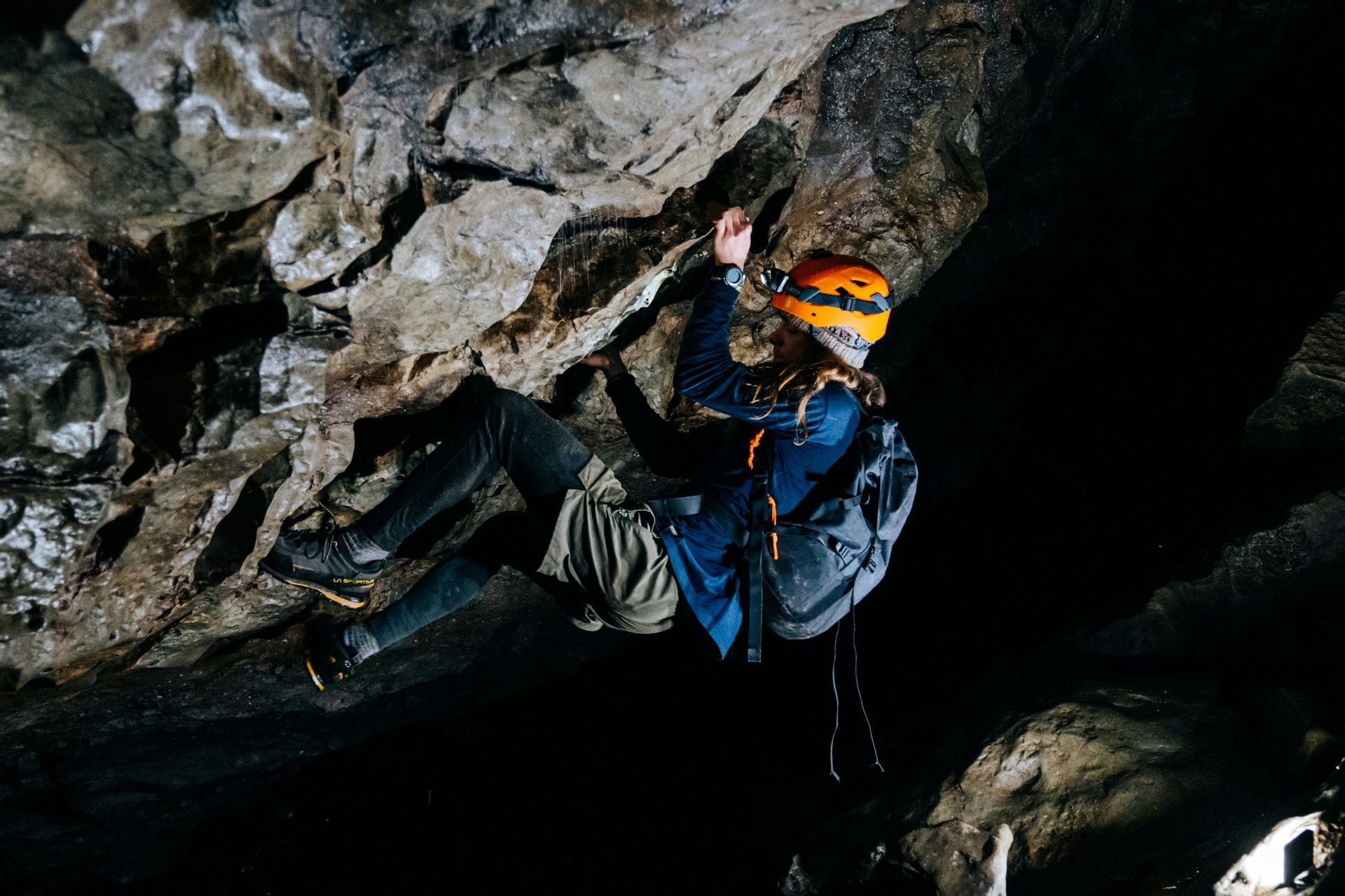 Una persona con un casco naranja y una linterna frontal escala la pared rocosa dentro de una cueva oscura.