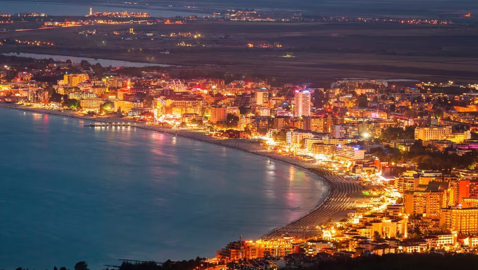 Vista aerea di una città costiera con edifici luminosi che fiancheggiano una spiaggia curva di notte.