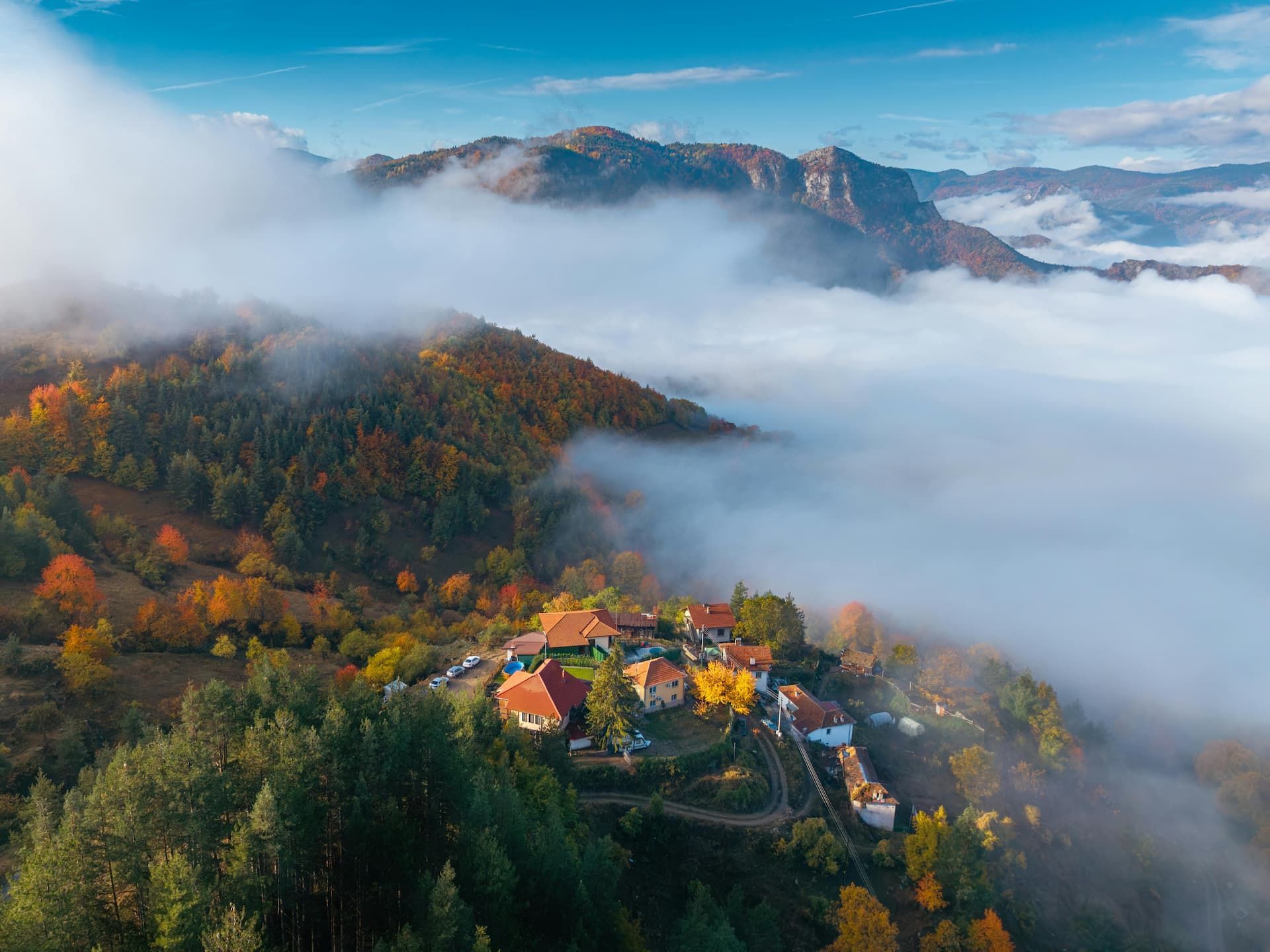 Vue aérienne d'un petit village à flanc de montagne, couvert d'arbres aux couleurs automnales, émergeant d'une mer de nuages.