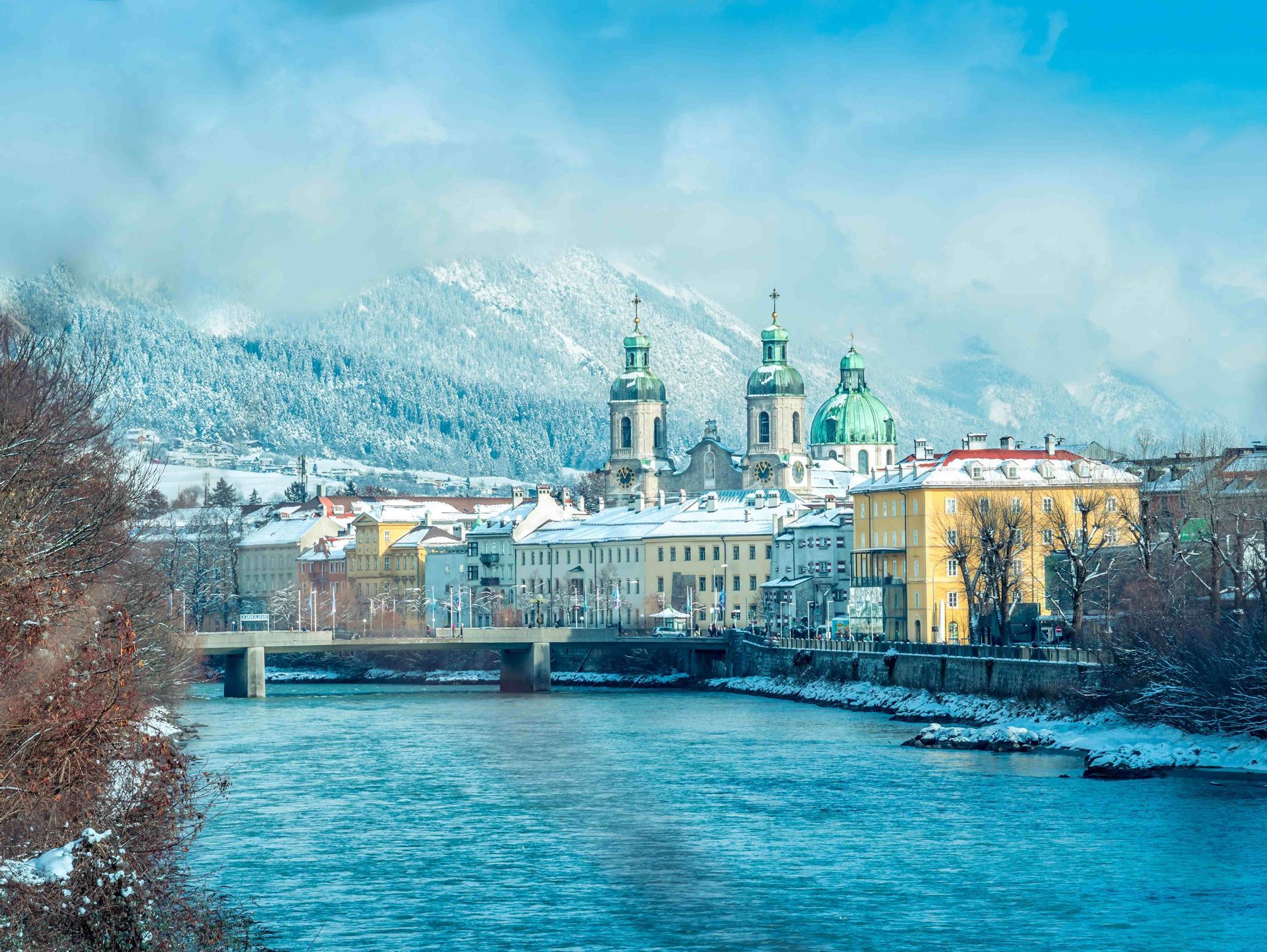 Ein Blick auf eine historische Stadt mit farbenfrohen Gebäuden und grünen Kirchtürmen entlang eines Flusses, mit großen schneebedeckten Bergen im Hintergrund.