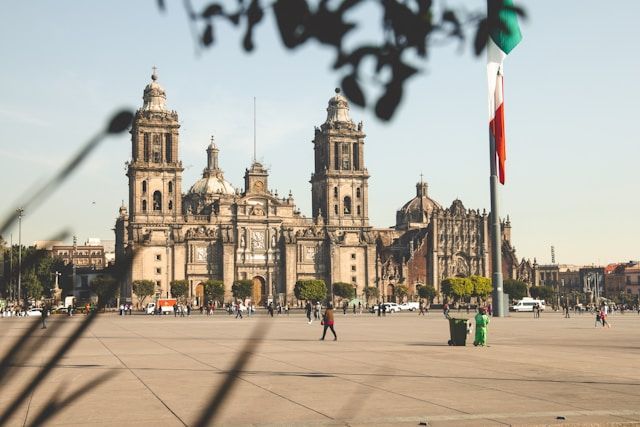 Une grande cathédrale historique avec des clochers jumeaux se dresse sur une place ensoleillée où les gens se promènent, avec un drapeau mexicain flottant à droite.