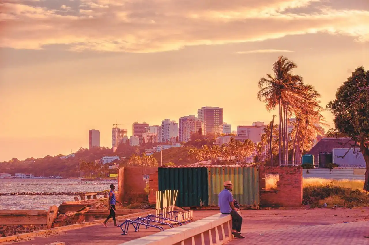 A man sits on a concrete ledge on a waterfront, looking out at a modern city skyline with palm trees during a warm sunset.