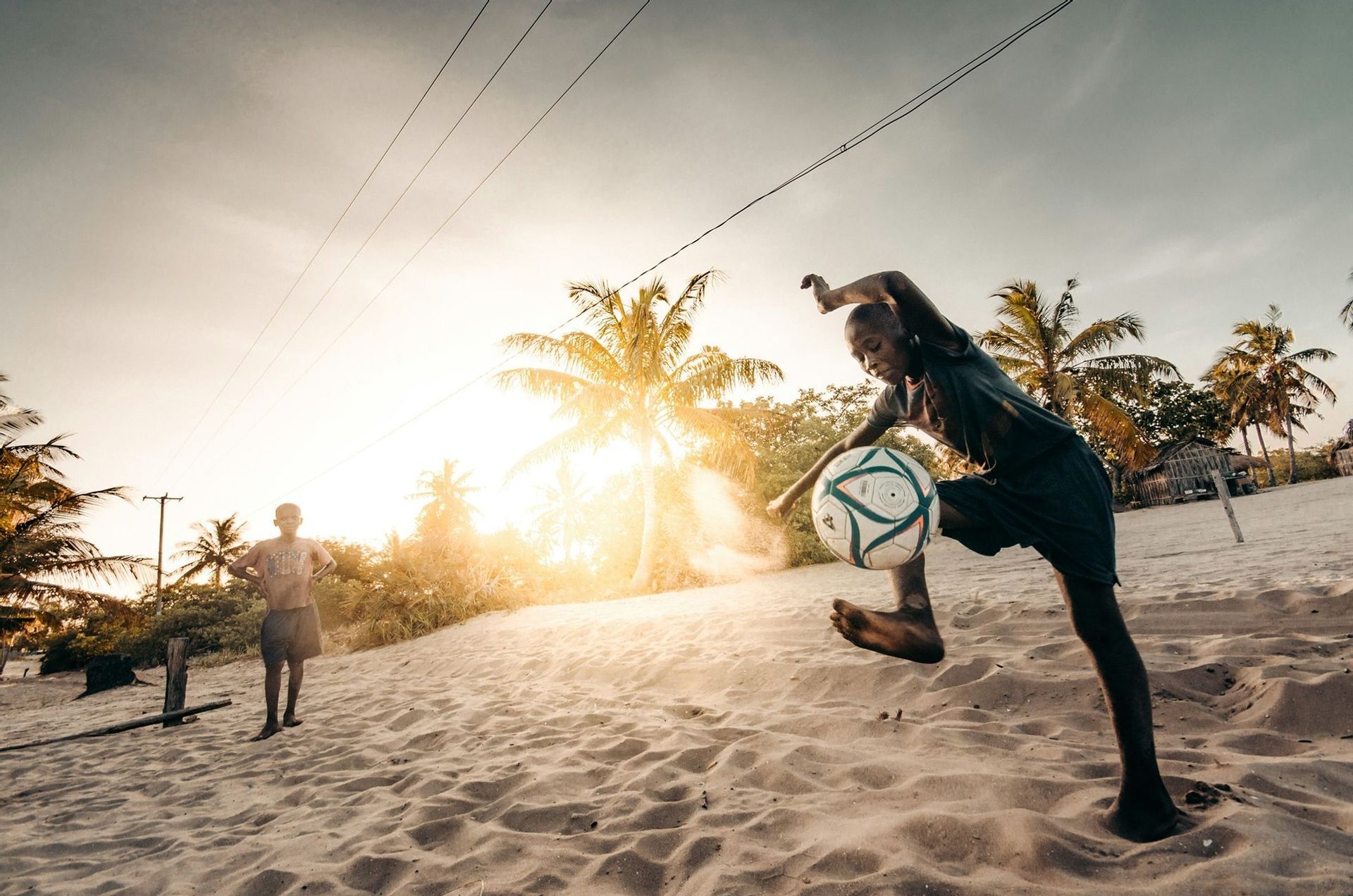 A young boy kicks a football in the air on a sandy beach at sunset, with another boy and palm trees in the background.