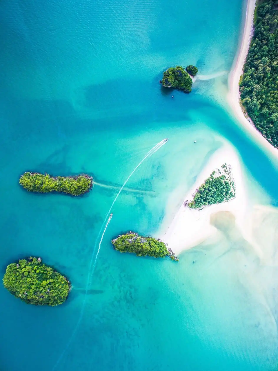 An aerial top-down view of motorboats creating wakes in turquoise water between small, lush green islands and white sandbanks.