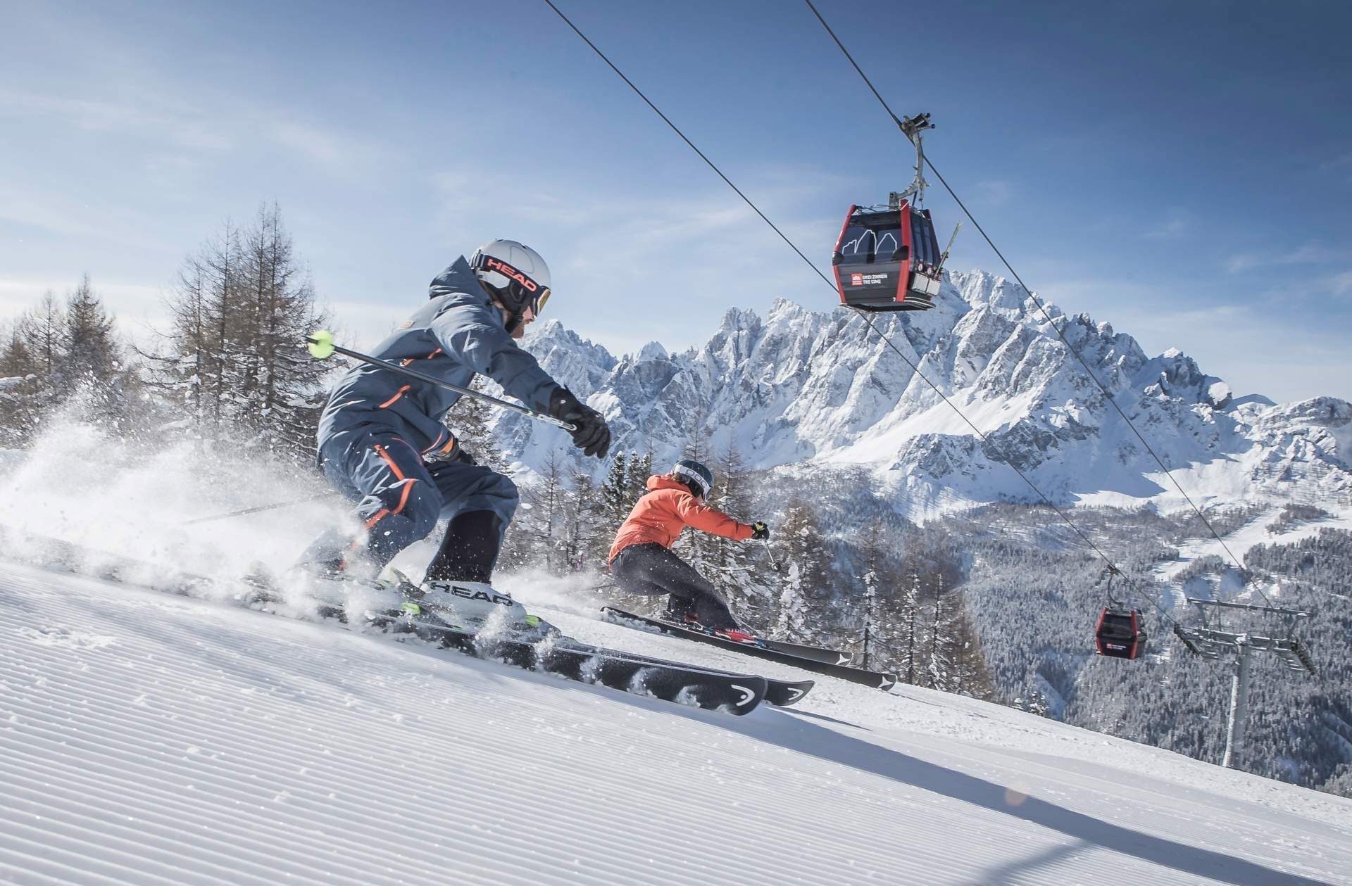 Two people from a WeRoad group trip skiing down a snowy slope, with a cable car overhead and mountains in the background.