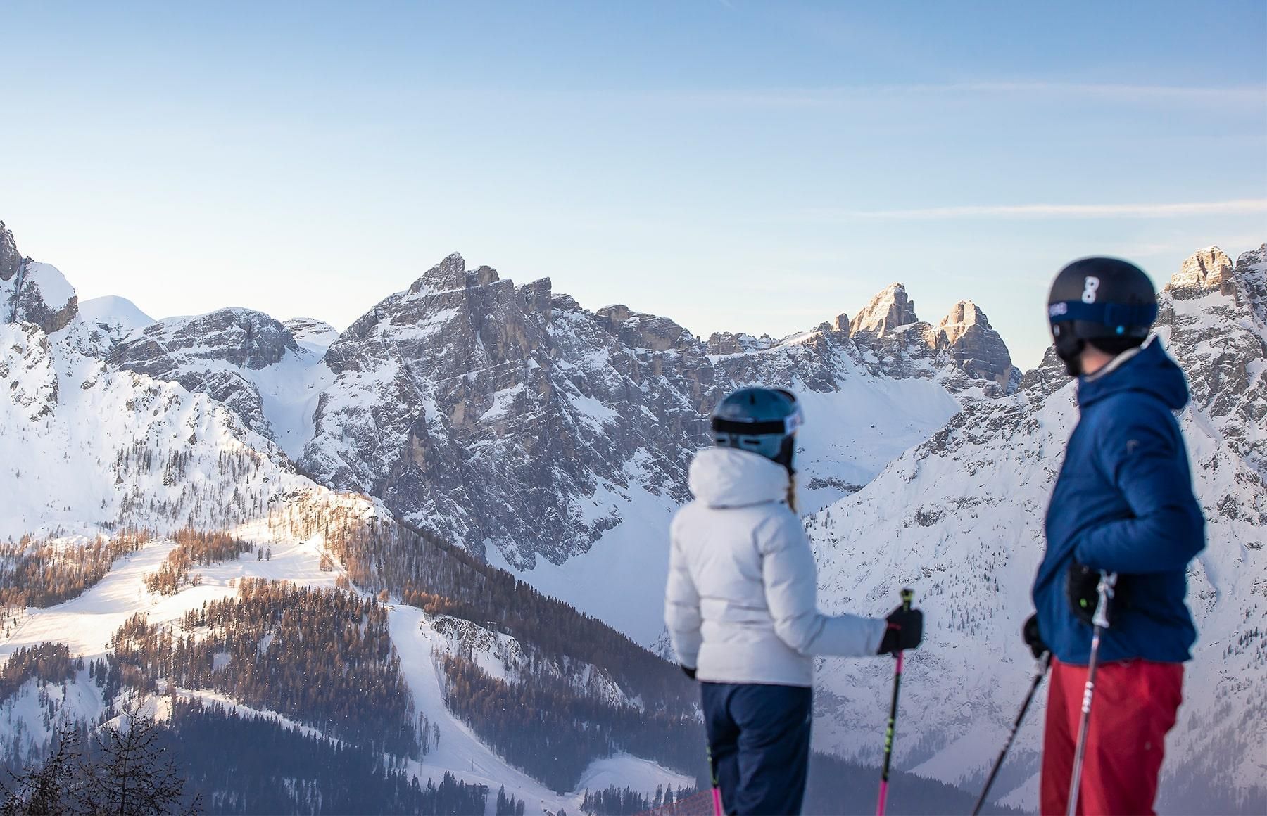 From behind, two skiers from a WeRoad group trip pause to look at a sunlit, snow-covered mountain range.