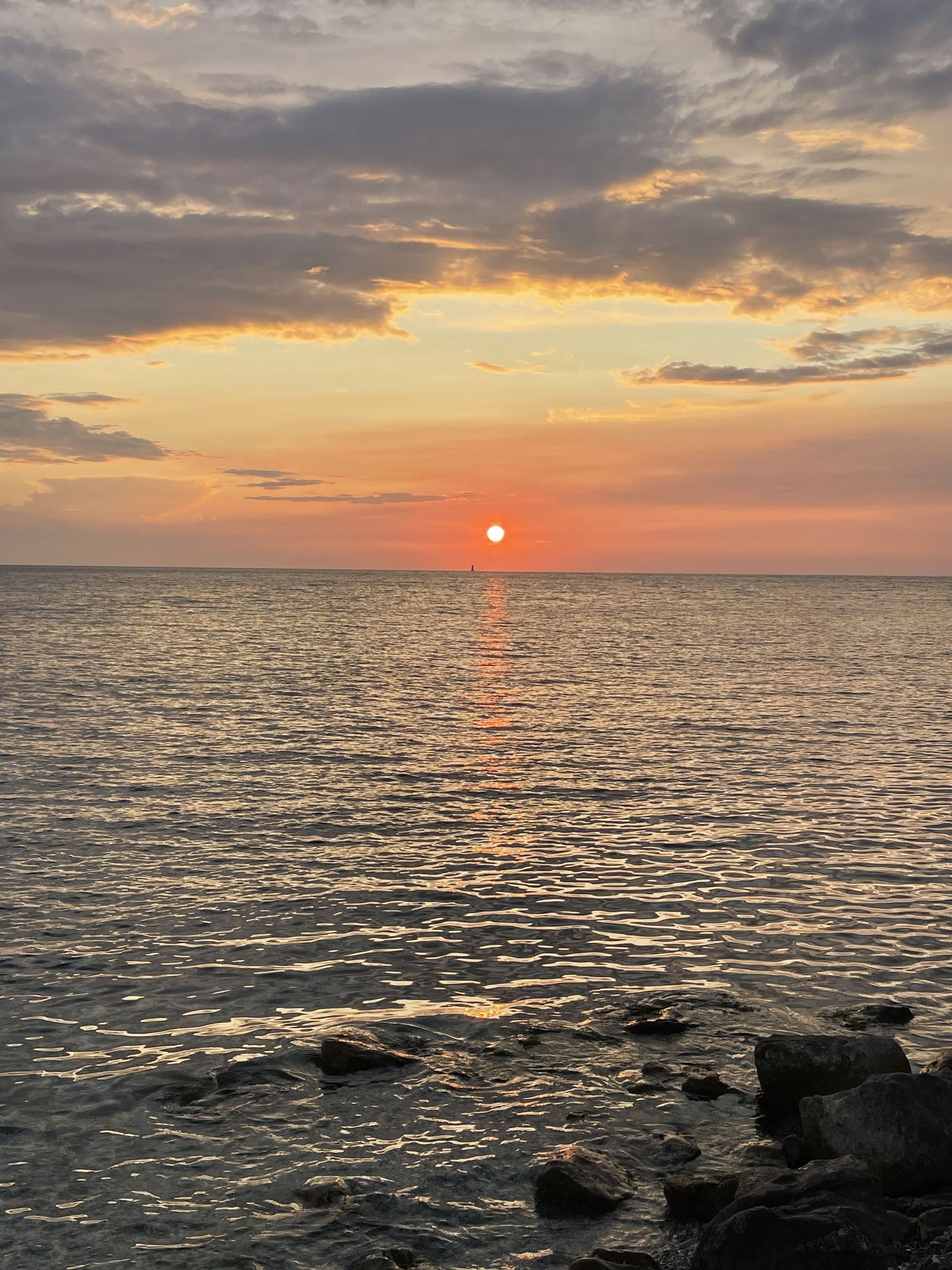 Il sole tramonta all'orizzonte sull'acqua, tingendo le nuvole di un bagliore arancione e riflettendosi sulla superficie increspata vicino a una riva rocciosa.
