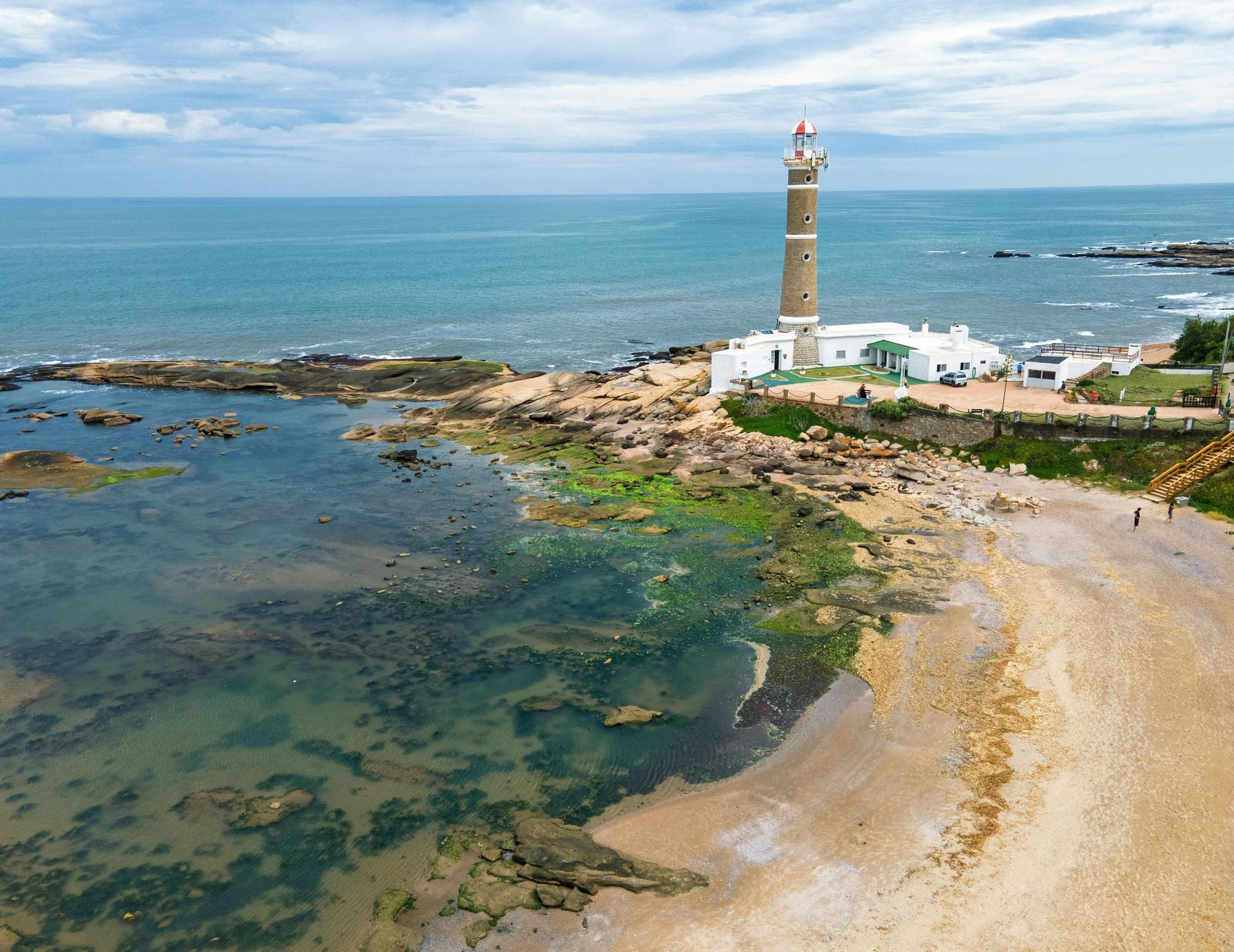 Veduta aerea di un alto faro in pietra che si erge su una costa rocciosa accanto a una caletta sabbiosa con acqua limpida.