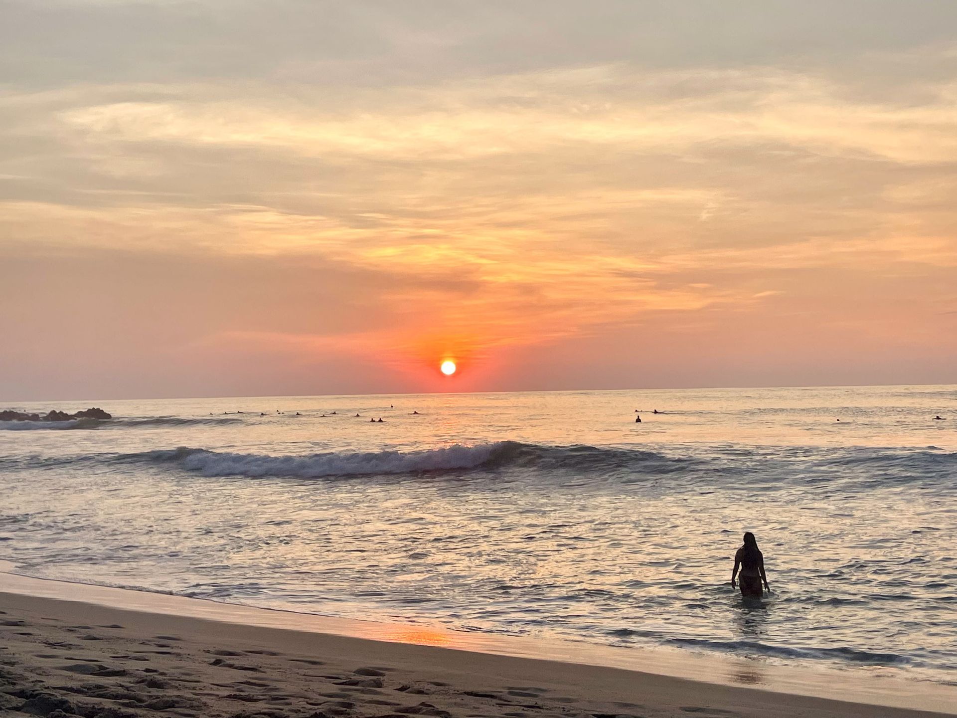 Une personne se tient dans l'eau peu profonde de l'océan au coucher du soleil, tandis que plusieurs surfeurs attendent les vagues au loin.