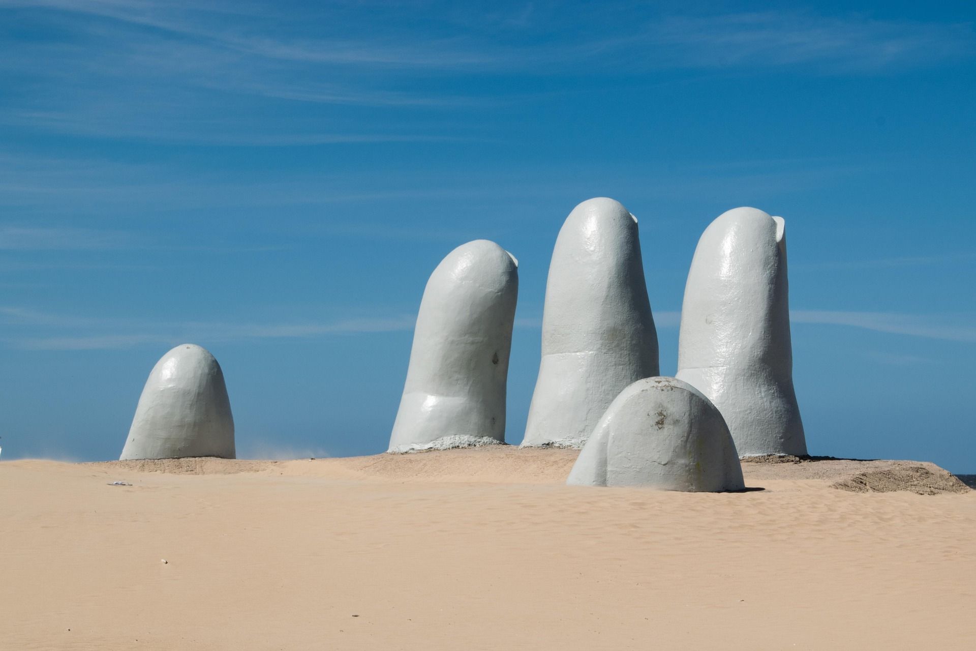 Grande scultura bianca di dita che affiorano dalla sabbia della spiaggia sotto un cielo azzurro.