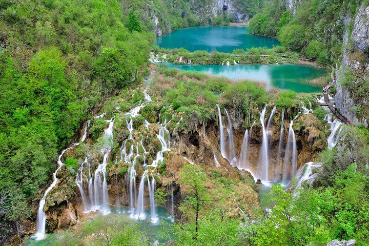 A landscape view of numerous waterfalls cascading down mossy cliffs into terraced turquoise lakes, all surrounded by dense green forest.