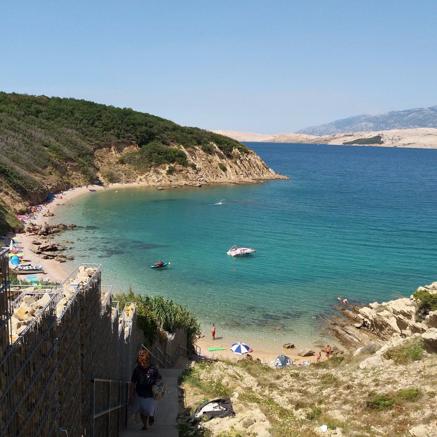 A high-angle view of a sunny coastal cove with turquoise water, a sandy beach with people, and boats floating near the shore.