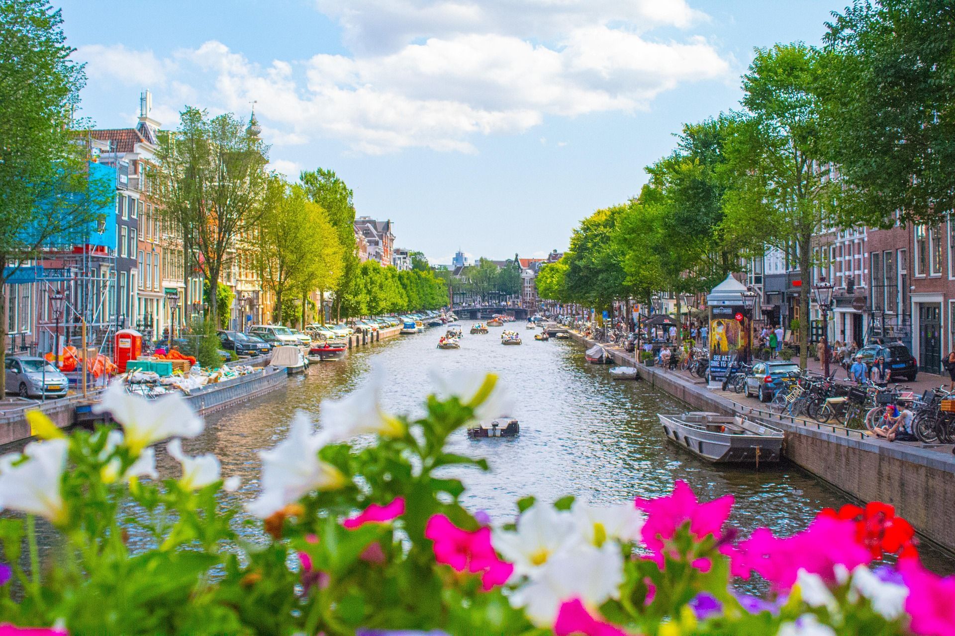 Vista di un canale cittadino soleggiato, con barche, edifici tradizionali e alberi verdi, e fiori colorati in primo piano.