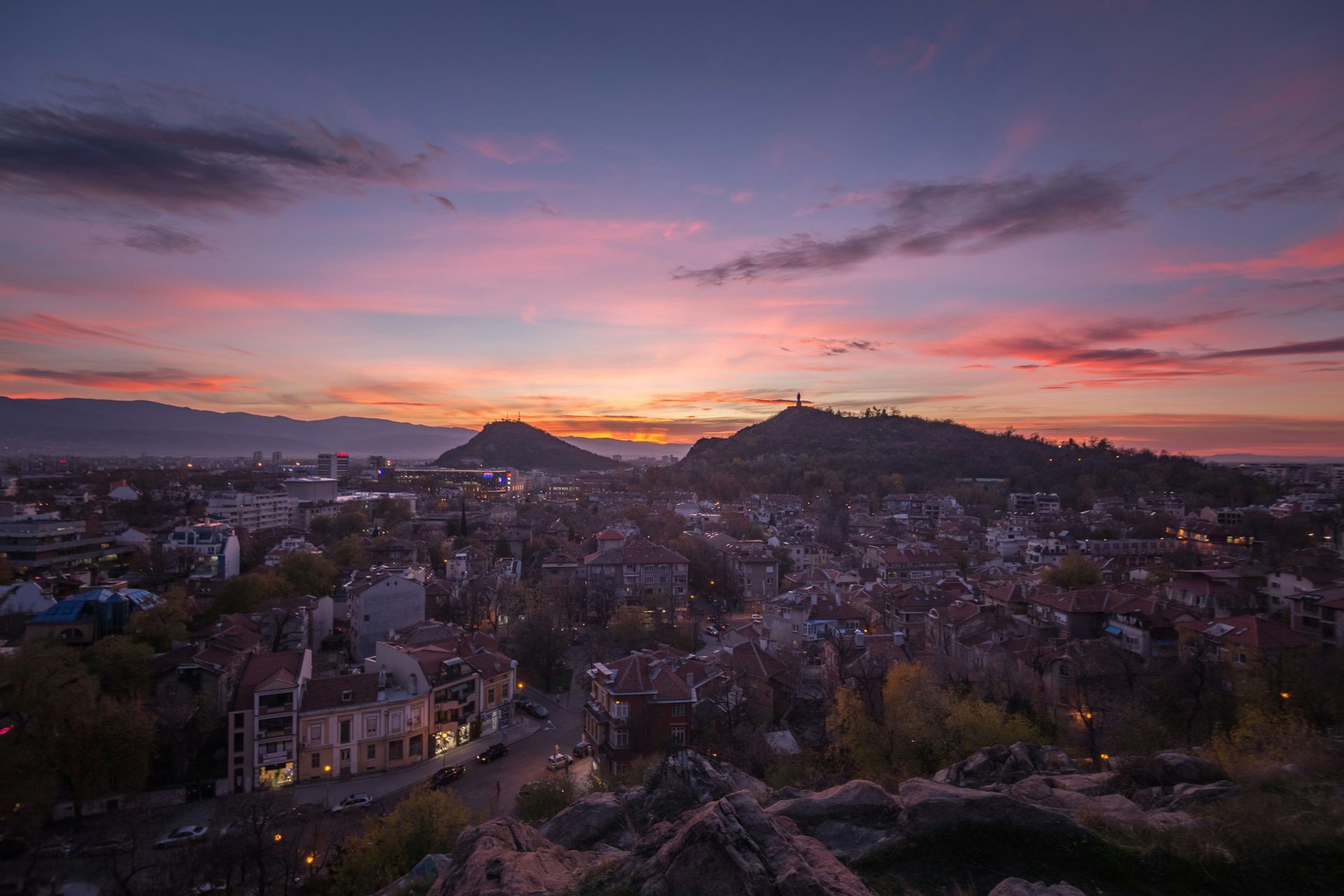 Une vue aérienne d'un paysage urbain niché entre deux grandes collines sous un ciel de coucher de soleil vibrant, rose, violet et orange.