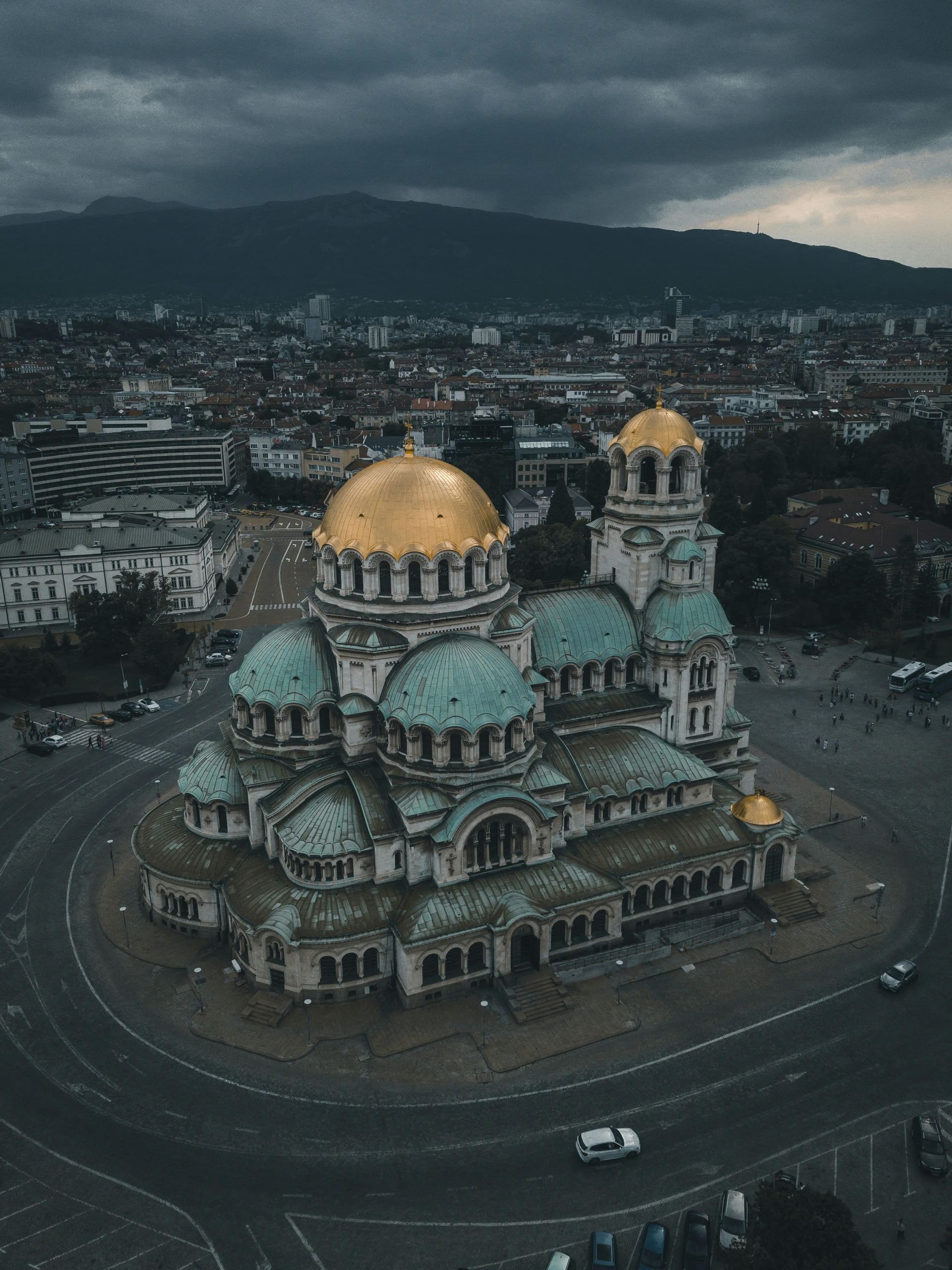 Une vue aérienne d'une grande cathédrale avec des dômes or et verts, entourée d'un paysage urbain, avec des montagnes sous un ciel nuageux.