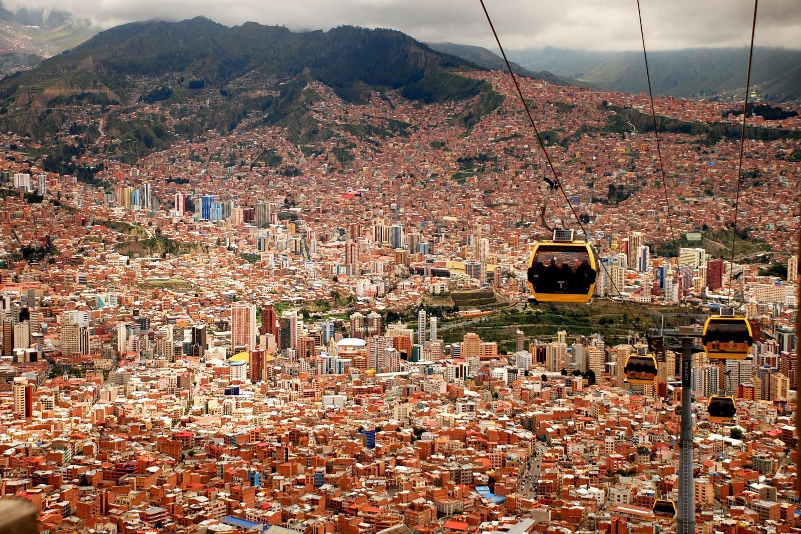 Yellow cable cars move over a dense cityscape of terracotta buildings spread across a valley at the base of a large mountain.