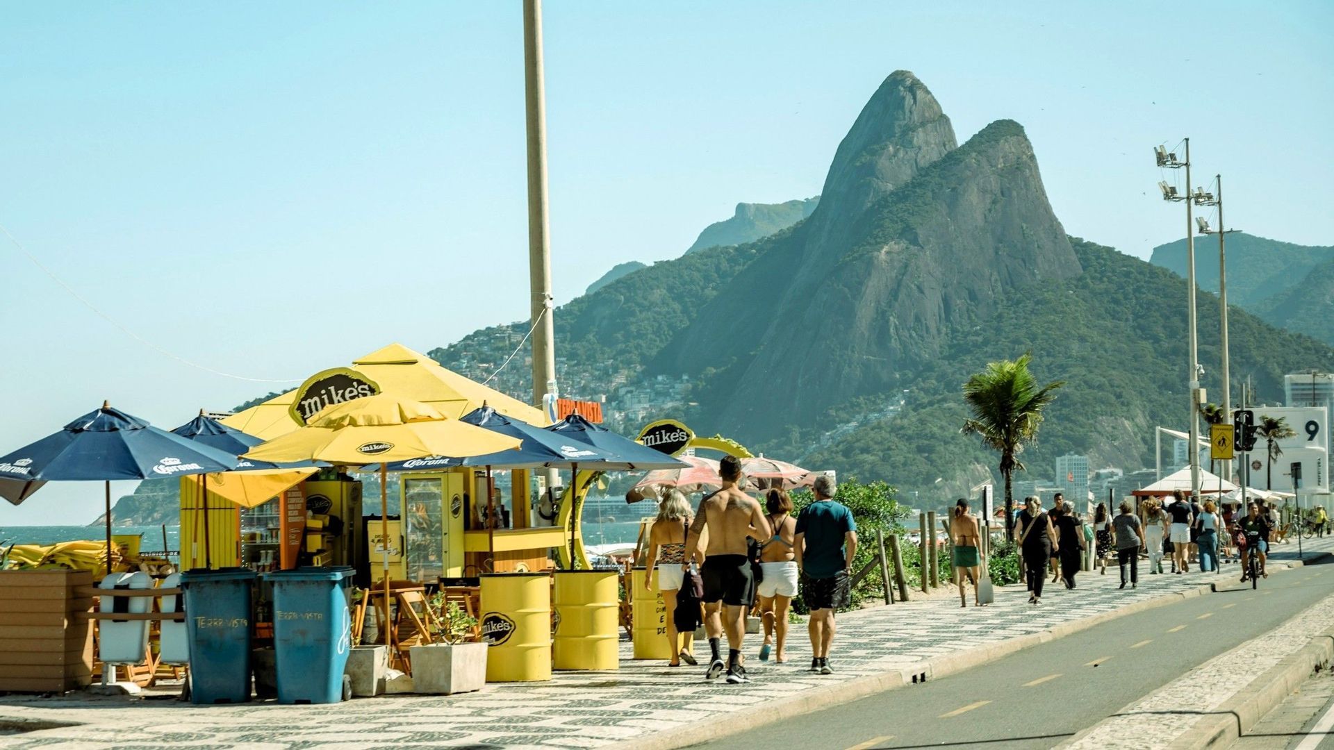 Menschen in Sommerkleidung spazieren auf einer gemusterten Strandpromenade an einem gelben Kiosk vorbei, im Hintergrund erheben sich große grüne Berge unter einem klaren Himmel.