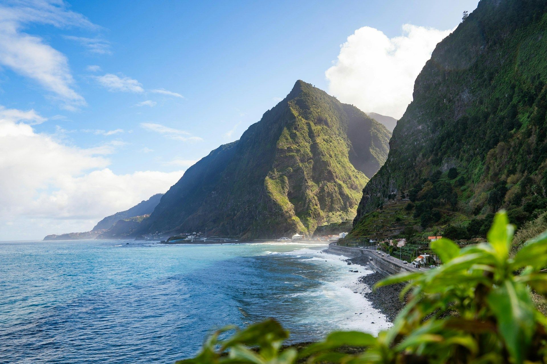 Una strada costiera si snoda lungo un litorale con onde turchesi, ai piedi di montagne ripide e verdi sotto un cielo parzialmente nuvoloso.