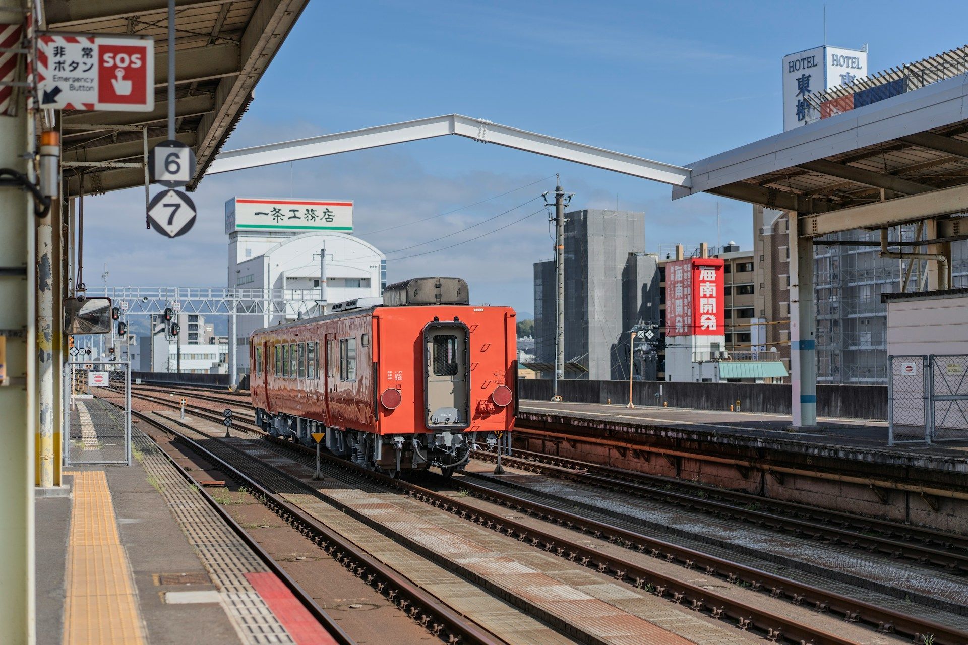 Un treno arancione sui binari su una banchina di stazione all'aperto con edifici cittadini visibili sullo sfondo sotto un cielo azzurro.