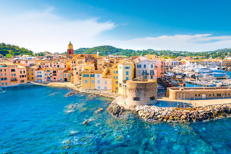 Una vista aerea di una colorata città costiera con una torre in pietra, un porto pieno di yacht e acqua turchese cristallina.
