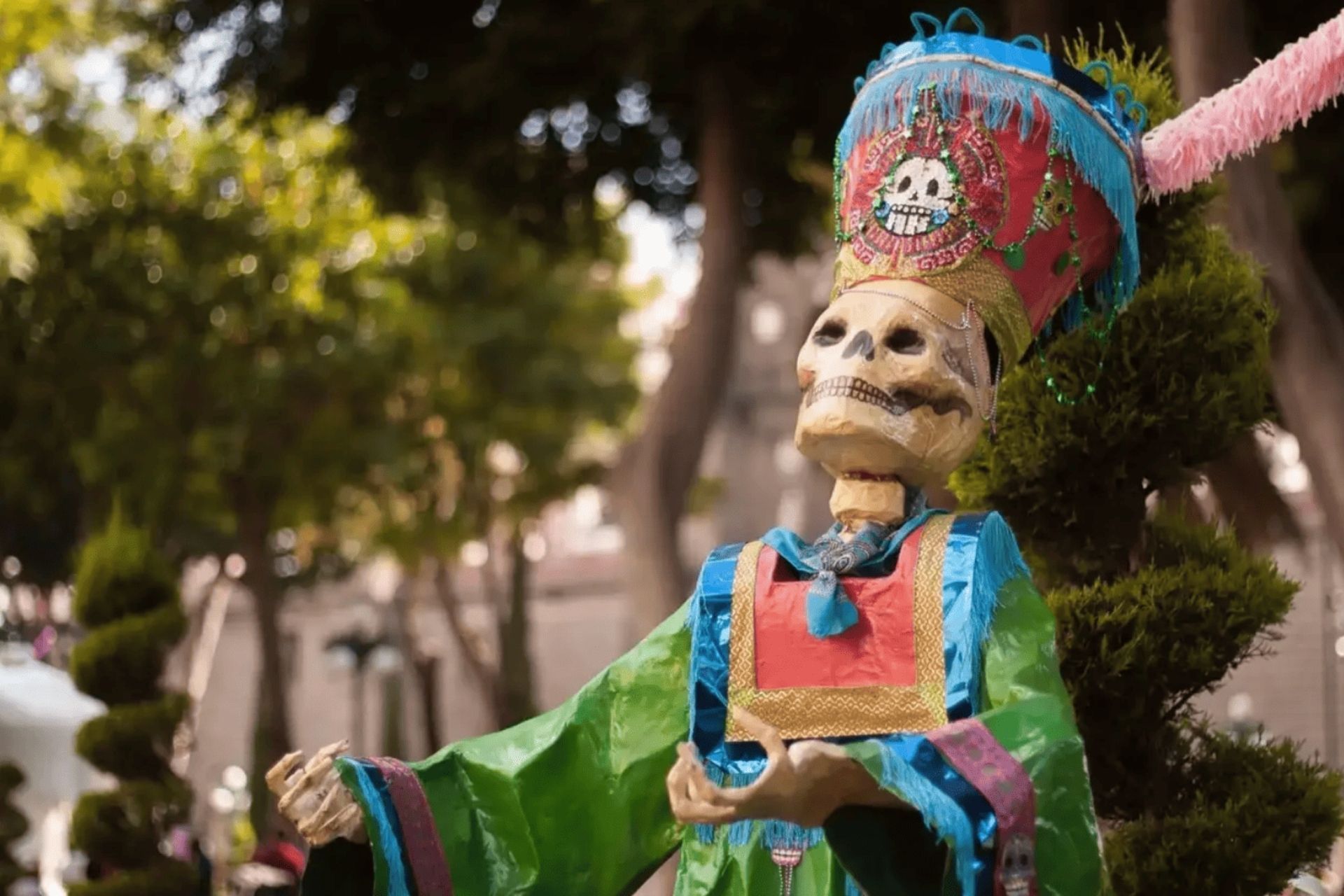Un squelette décoratif vêtu d'un costume coloré et d'une coiffe haute et élaborée ornée d'un emblème de crâne, se dresse dans un parc.