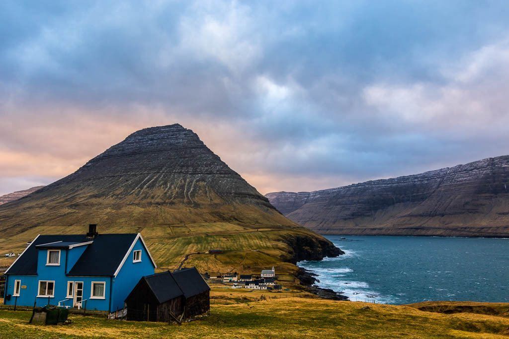 Una casa blu su una collina erbosa si affaccia su uno specchio d'acqua e una grande montagna sotto un cielo nuvoloso.